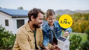 Junger Mann steht mit seiner Tochter im Garten mit einem Modellhaus in der Hand und zeigt auf eine Photovoltaik-Anlage auf dem Dach, Modernisierungskredit ab 2,79% p.a.*