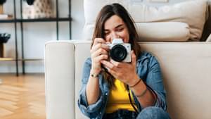 Eine Frau sitzt angelehnt vor dem Sofa und hält eine Kamera vor sich und schaut lächelnd in die Anzeige der gemachten Aufnahme der Kamera.