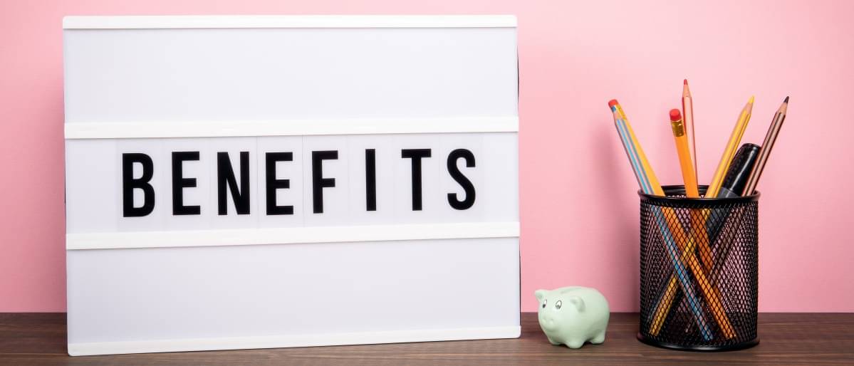 On a table there is an illuminated board with the words "Benefits". Next to it is a cup with pens and a piggy bank. 