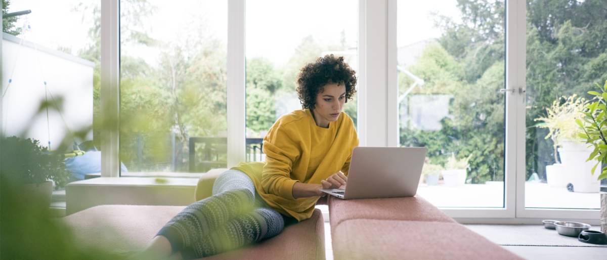 Eine junge, dunkelhaarige Frau sitz auf einem Sofa, arbeitet entspannt am Laptop.