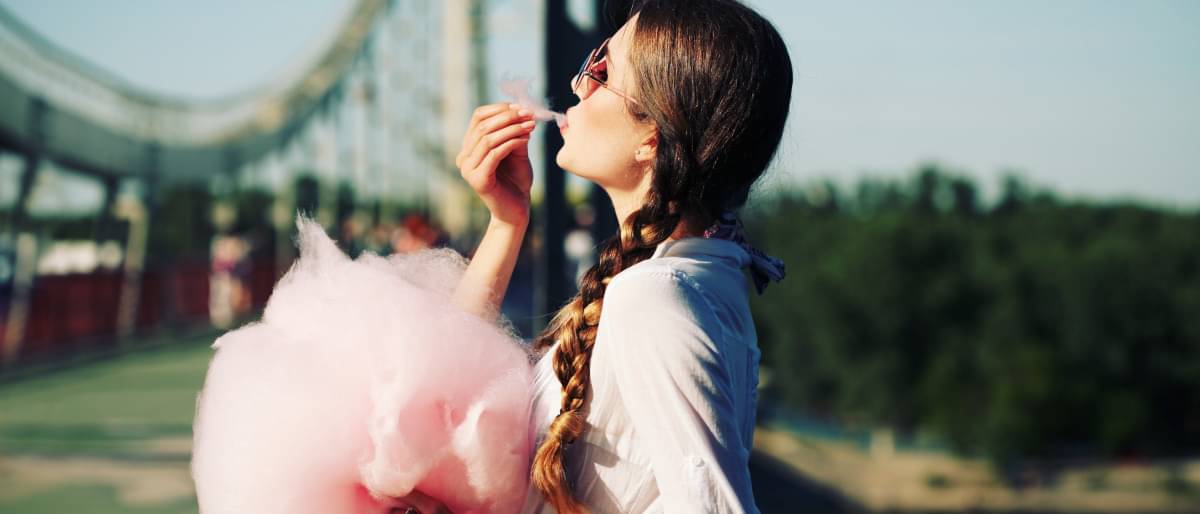 Junge Frau mit Zöpfen steht an einem Brückengeländer lehnend und isst rosa Zuckerwatte