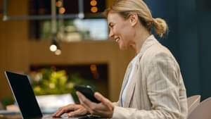 Frau im Business-Outfit sitzt lächelnd an einem Schreibtisch, hält ein Smartphone in der Hand und arbeitet gleichzeitig an einem Laptop in moderner Umgebung.