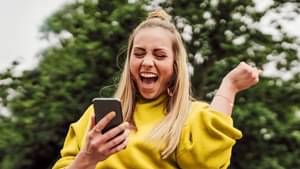 Eine junge, blonde Frau mit Zopf und gelben Pulli schaut auf ihr Handy und freut sich und hebt dabei einen Arm mit der Hand zu Faust geballt nach oben.