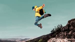 Ein Mann mit gelber Jacke und Rucksack springt von einem Felsabsatz herunter und streckt beide Arme zur Seite aus.