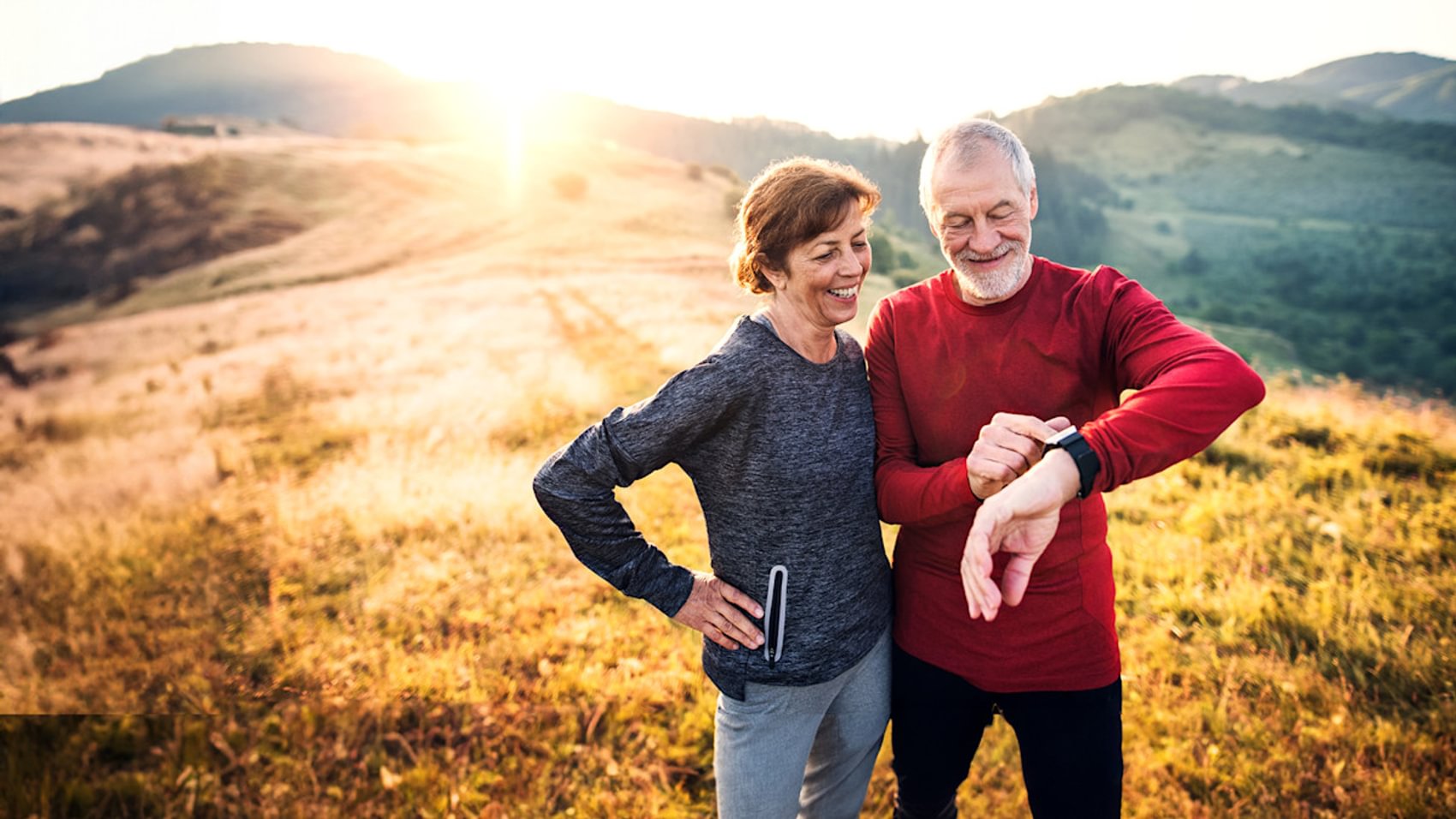 Eine Frau und ein Mann sind im hügeligen Gelände beim Running unterwegs und machen gerade eine Pause und schauen dabei auf Ihre Pulsuhr.