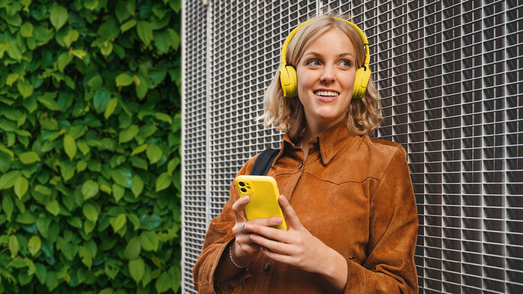 Frau hält Handy in der Hand und hört über Kopfhörer Musik und lächelt mit einem Blick leicht zu Seite gerichtet.