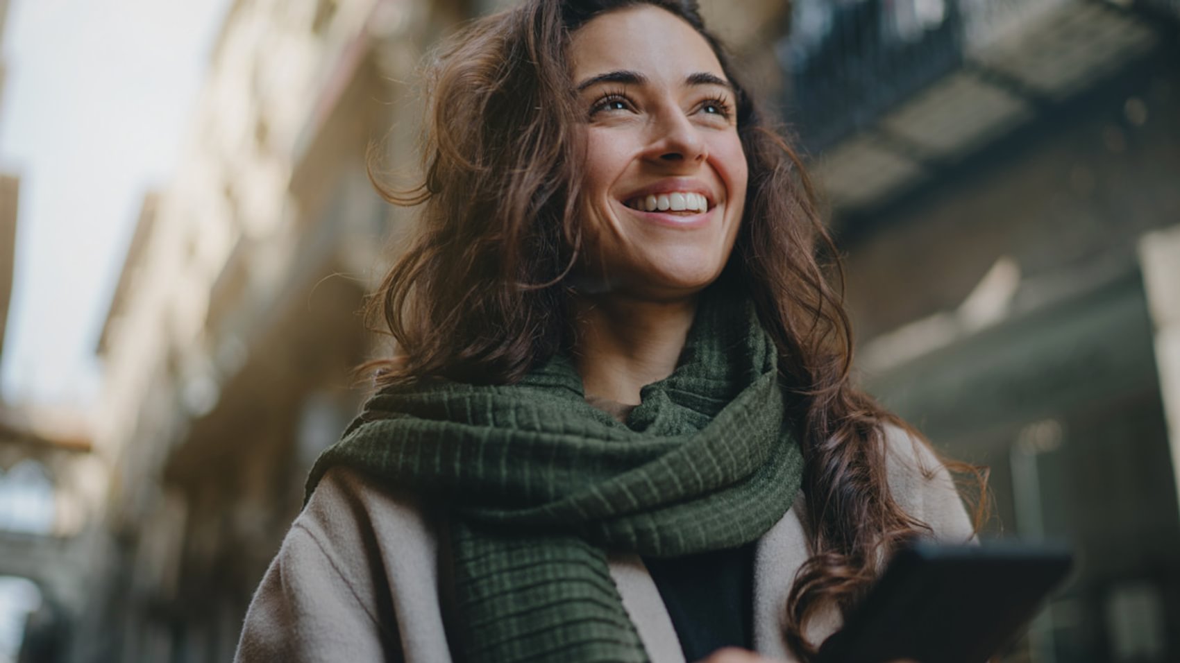 Eine junge, attraktive Frau mit einem grünen Schal lächelt, während sie an einem kalten Tag durch die Straßen einer Stadt läuft und auf ihr Smartphone schaut. 