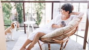 Junge Frau sitzt in einem Korbstuhl auf dem Balkon zusammen mit ihrem Hund.