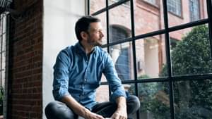 Mann mit kurzen dunklen Haaren und Bart sitzt in Fensterrahmen und schaut nach draußen.