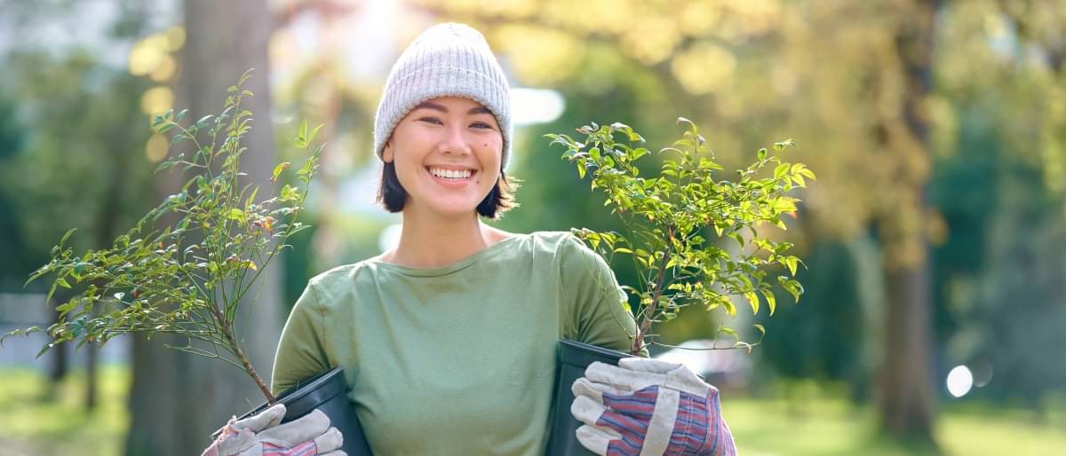 A woman wearing a hat and gardening gloves stands in a park and holds two small tree saplings in her arms. She smiles at the camera.