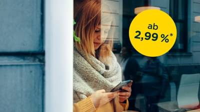 Junge Frau steht hinter bodentiefen Fenster mit Schal und gelbem Oberteil. Sie hält Handy in der Hand und tippt auf dem Bildschirm: Onlinekredit ab 2,99% p.a.*