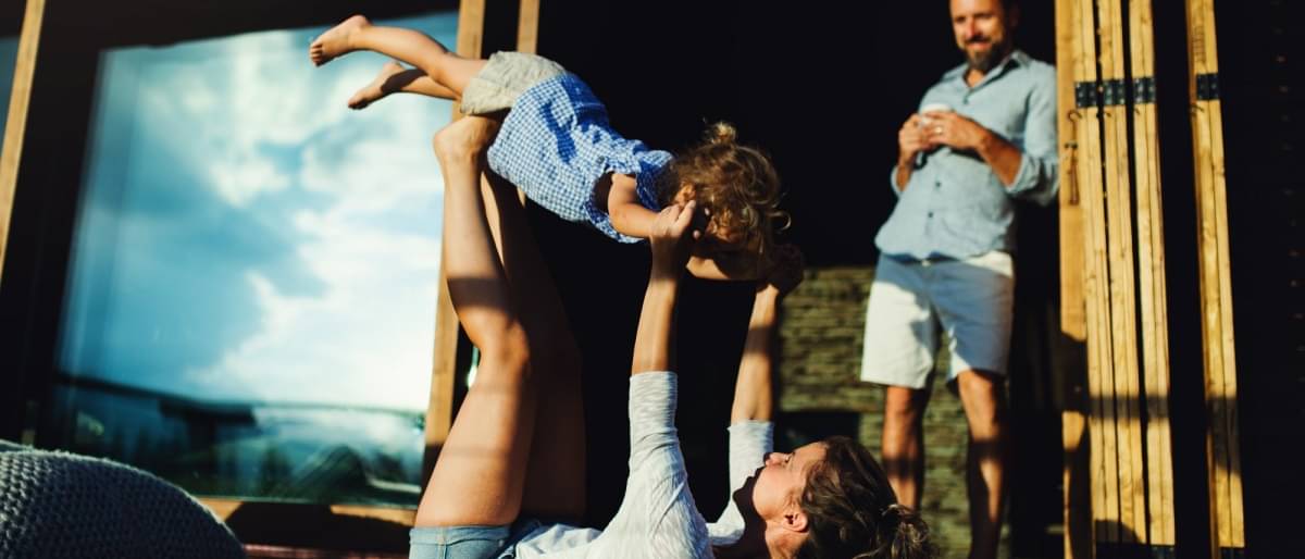 Eine Mama spielt mit ihrer Tochter auf der Terasse und macht Turnübungen. Ihr Mann steht lachend in der Tür und schaut den beiden amüsiert zu.