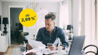 Junger Mann sitzt an Tisch vor Laptop und hat Unterlagen und Handy in den Händen: Umschulung ab 2,99%*