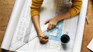 Eine Frau sitzt an einem Tisch und fertigt mit Lineal und Geodreieck eine Bauzeichnung an.