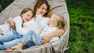 Eine Mutter liegt mit ihren zwei kleinen Töchtern entspannt in einer Hängematte im Garten. Sie hält beide Mädchen beschützend im Arm, die sich an ihre Mutter kuscheln.