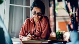 Frau mit dunklen Haaren im Pferdeschwanz gebunden und Brille sitzt an Tisch und schreibt auf Papierunterlagen.