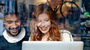 Dunkelhäutiger Mann mit Bart und Frei mit roten langen Haaren sitzen vor Laptop und lachen.