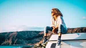 Frau mit Locken sitzt glücklich auf ihrem Camper-Van und genießt die Aussicht